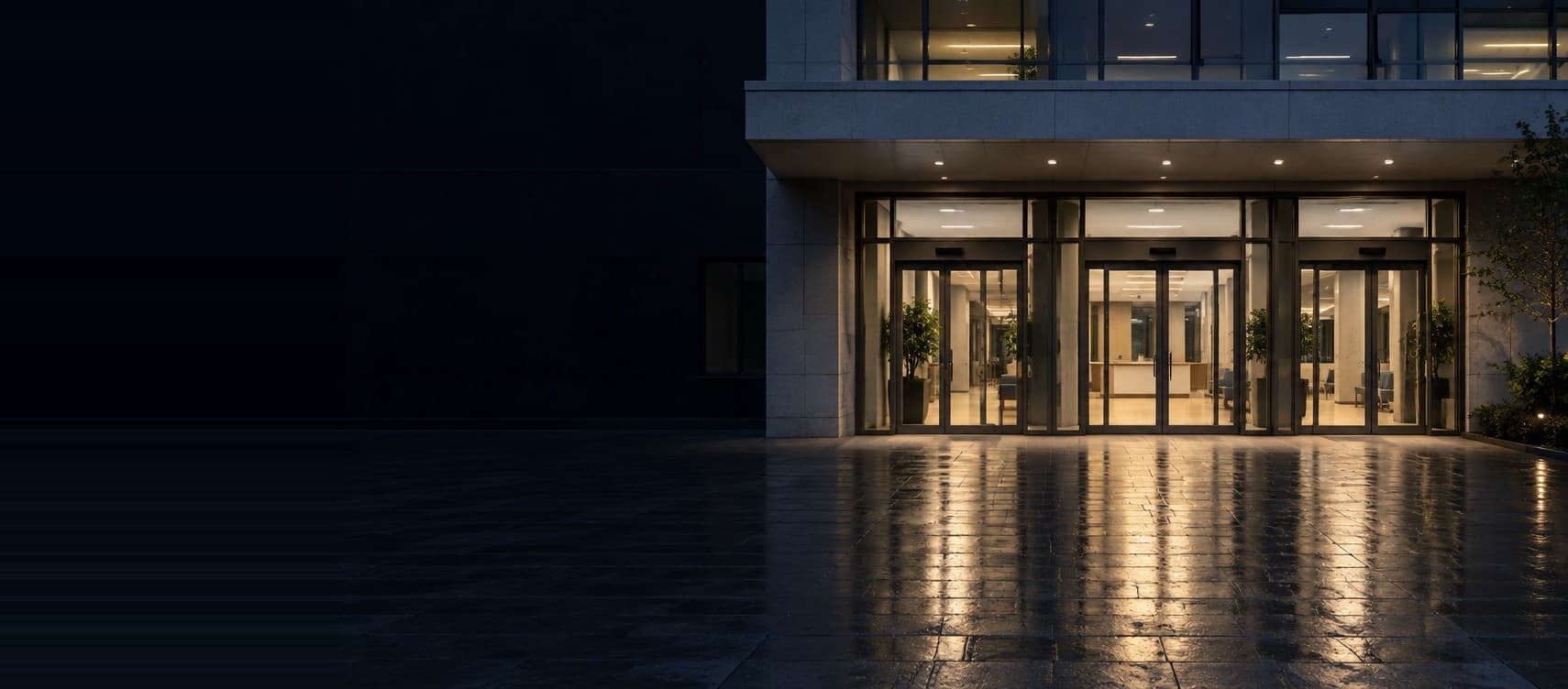 A hospital entrance at dusk, three glass doors lit warm from the lobby.
