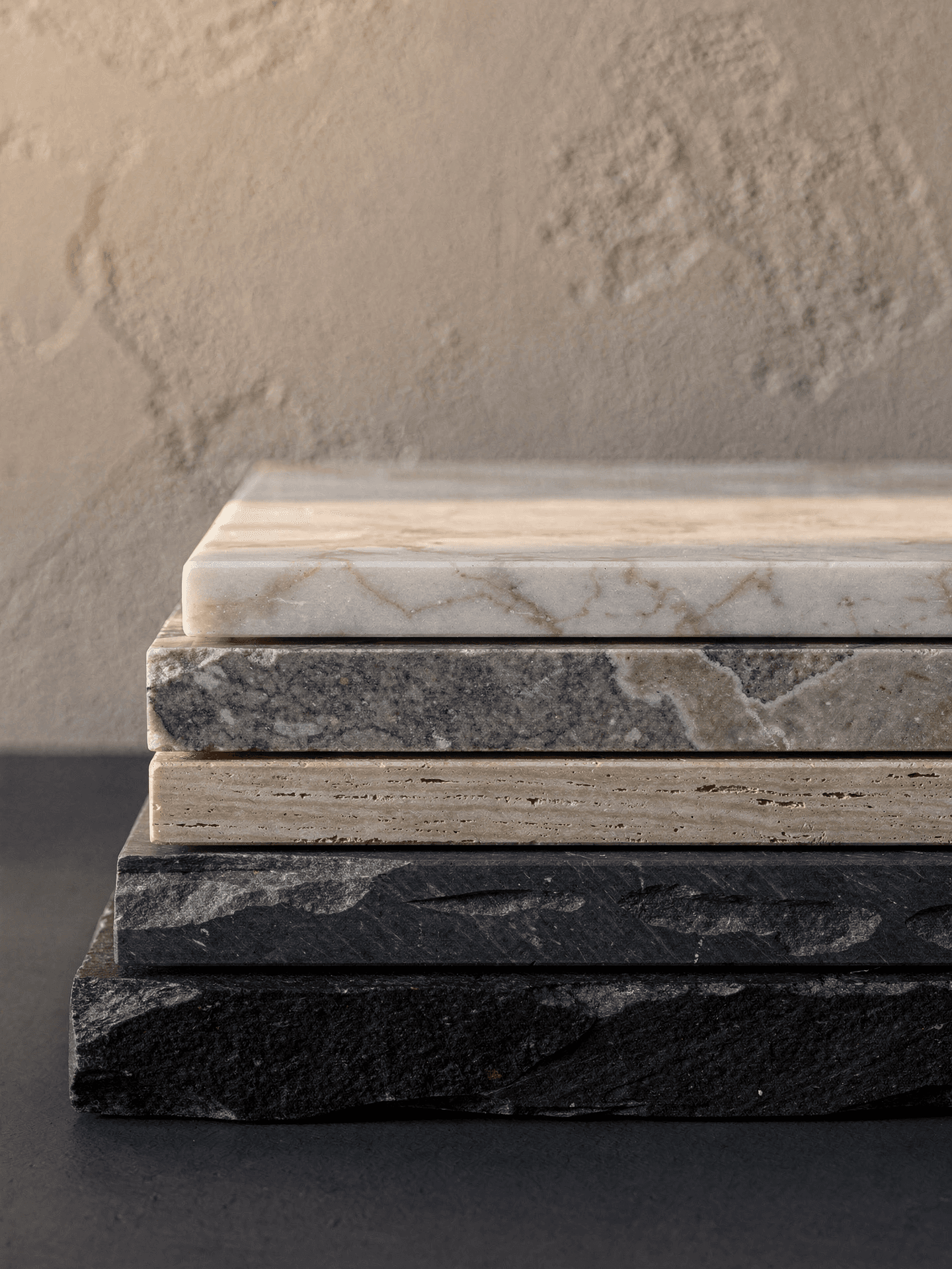 Five slabs of stone stacked on a dark surface against a plaster wall, dark slate at the base and pale marble at the top, lit from the left.