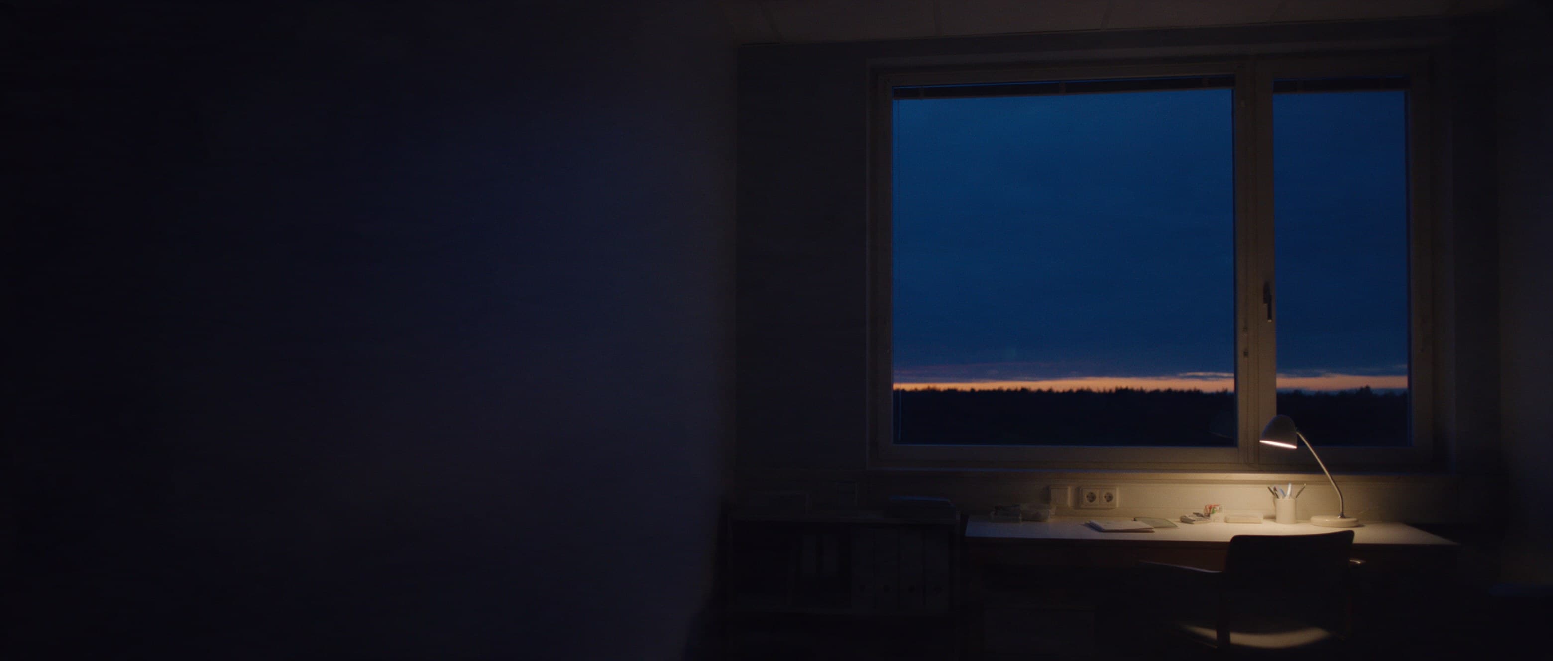 The room behind the lit window at dusk: a desk under one lamp, the evening horizon beyond, plain binders in shadow.